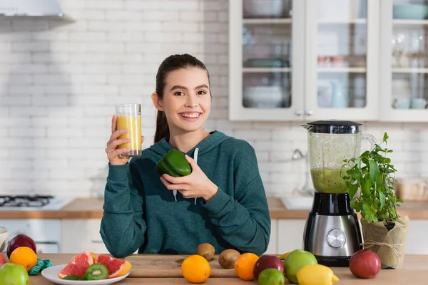 Mulher feliz segurando vidro de smoothie e pimentão perto de frutas frescas na cozinha — Fotografia de Stock