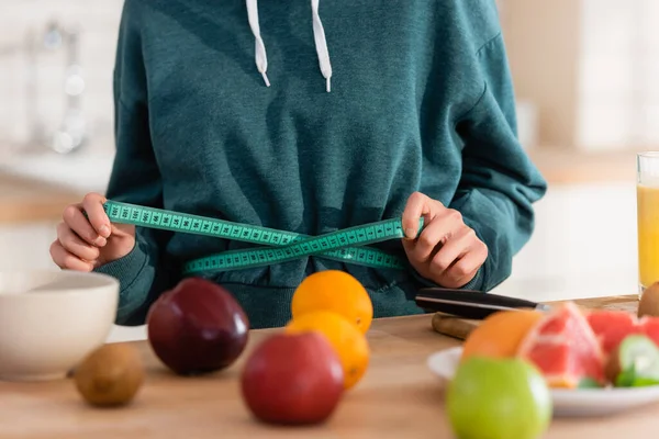 Cropped view of woman measuring waist near fresh fruits on blurred foreground — Stock Photo