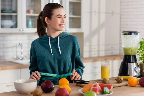 Donna gioiosa che distoglie lo sguardo mentre misura la vita vicino alla frutta fresca in cucina — Foto stock