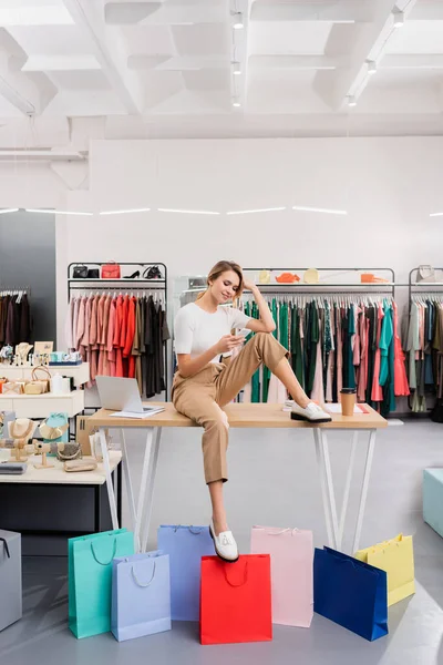 Vendedor sonriente usando teléfono inteligente cerca de bolsas de compras en la sala de exposición - foto de stock