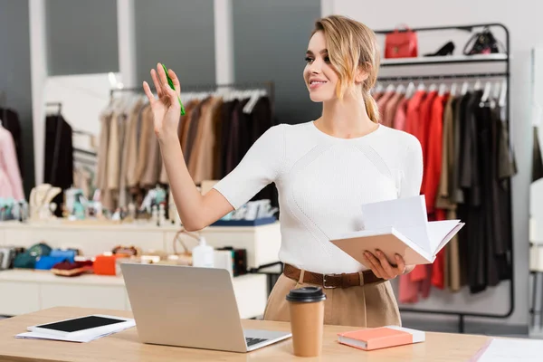 Smiling seller with notebook waving hand near devices in showroom — Stock Photo