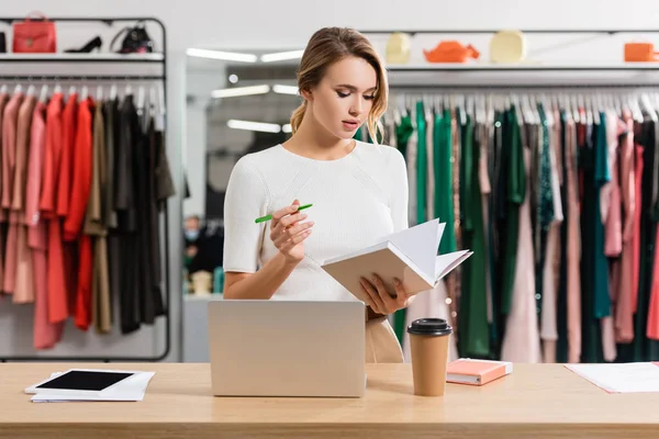 Seller working with pen and notebook near devices and paper cup in showroom — Stock Photo