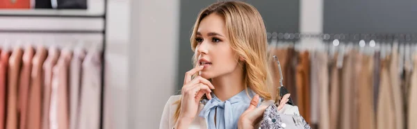 Pensive woman holding hanger with dress in showroom, banner — Stock Photo