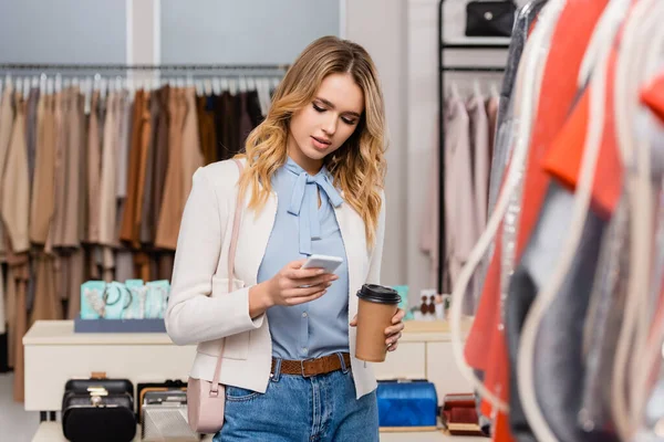 Mulher loira usando smartphone e segurando copo de papel perto de roupas no showroom — Fotografia de Stock