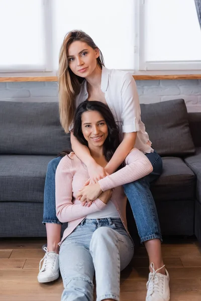 Lesbianas rubias y morenas felices abrazándose en la sala de estar - foto de stock