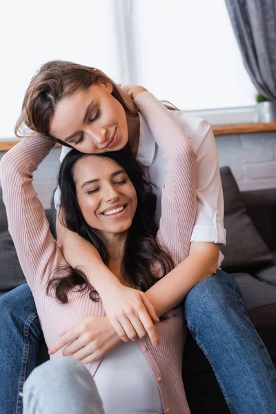 Lesbianas sonrientes con los ojos cerrados abrazándose en la sala de estar - foto de stock