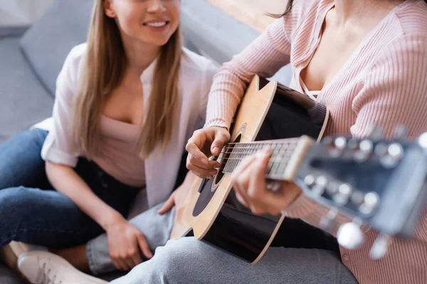 Vue recadrée de la femme jouant de la guitare acoustique près petite amie heureuse dans le salon — Photo de stock