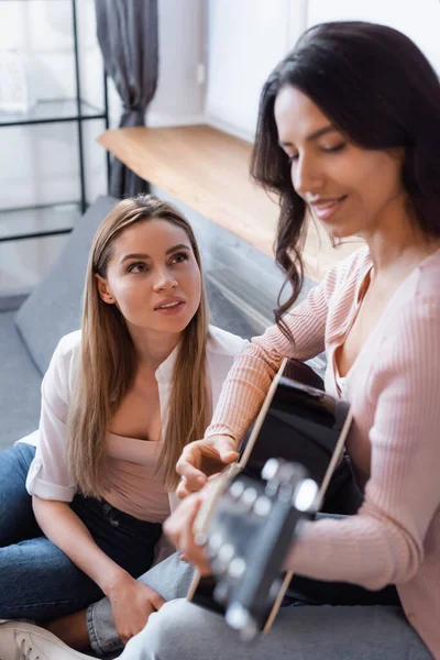 Blurred brunette woman playing acoustic guitar near girlfriend in living room — Stock Photo