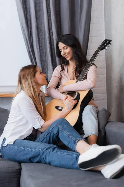 Femme brune et petite amie joyeuse tenant la main près de la guitare acoustique dans le salon — Photo de stock