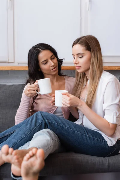 Girlfriends holding cups of tea while sitting on sofa in living room — Stock Photo
