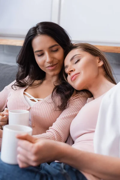 Happy girlfriends holding cups of tea while chilling on sofa in living room — Stock Photo