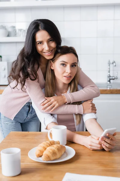 Happy woman hugging girlfriend with smartphone near breakfast on table — Photo de stock