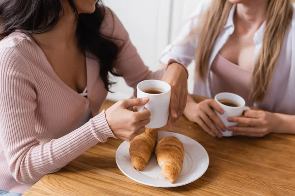 Abgeschnittene Ansicht von lesbischem Paar, das Tassen in der Nähe von Croissants auf dem Tisch hält — Stockfoto