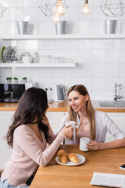 Happy lesbian couple holding cups near croissants on table — Stock Photo