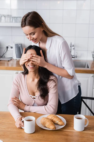 Happy woman covering eyes of cheerful girlfriend in kitchen — Photo de stock