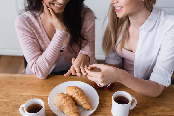 Cropped view of smiling woman wearing wedding ring on finger of cheerful girlfriend at home - foto de stock