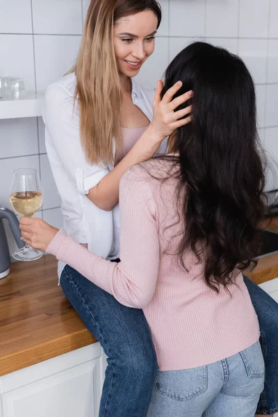 Happy woman sitting on kitchen table and looking at girlfriend with glass of wine - foto de stock