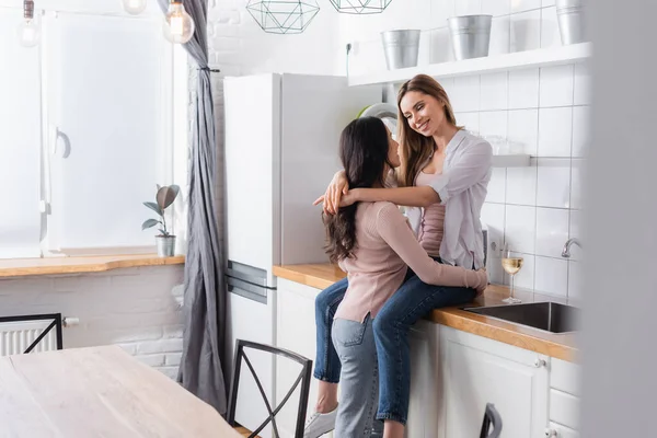 Happy woman sitting on kitchen table and hugging girlfriend near glass of wine — Fotografia de Stock