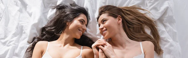 Top view of happy lesbian couple in white bras holding hands in bedroom, banner — Fotografia de Stock