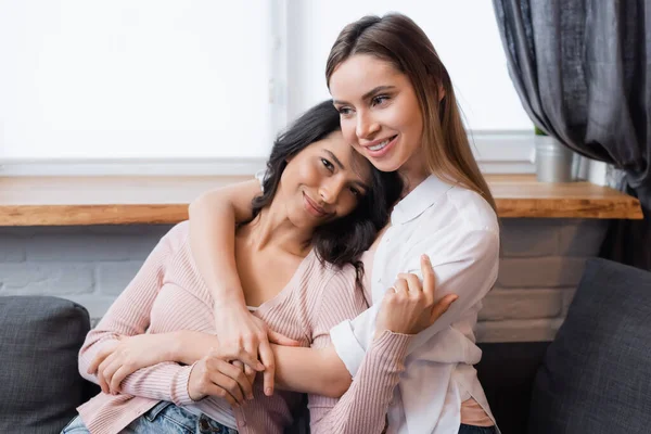 Smiling lesbian woman embracing happy girlfriend in living room - foto de stock