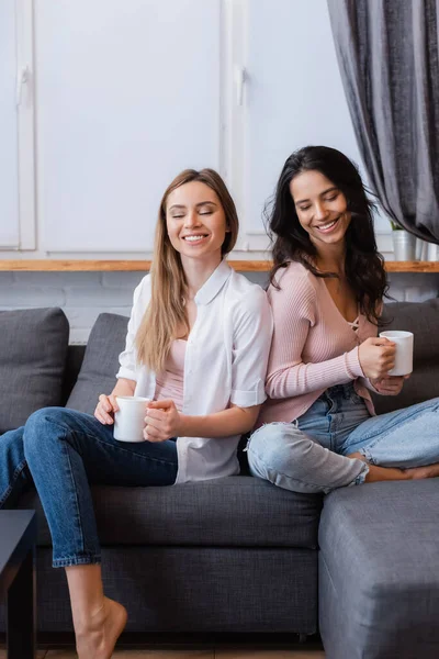 Pleased girlfriends holding cups of tea while chilling on sofa in living room — Stock Photo