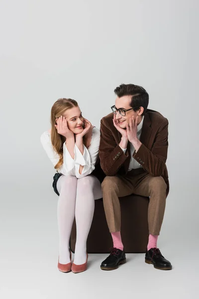 Jovem casal em roupas retro sorrindo um para o outro enquanto sentado em mala no fundo cinza — Fotografia de Stock