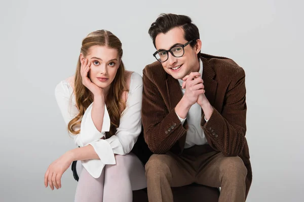 Homme et femme souriants en vêtements vintage regardant la caméra tout en étant assis sur une valise isolée sur gris — Photo de stock