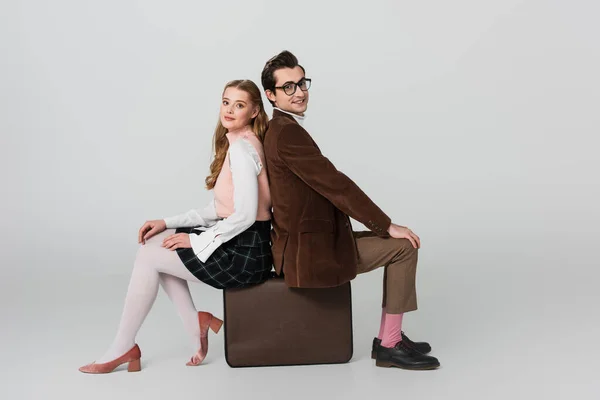 Joyful, retro style couple looking at camera while sitting back to back on suitcase on grey background — Stock Photo