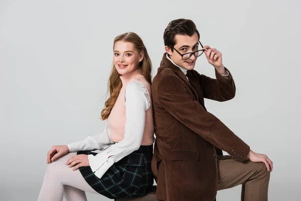 Smiling man touching eyeglasses while sitting on suitcase back to back with girlfriend isolated on grey — Stock Photo