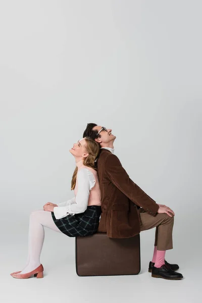 Glückliches Paar im Retro-Stil, Rücken an Rücken auf Koffer vor grauem Hintergrund sitzend — Stockfoto