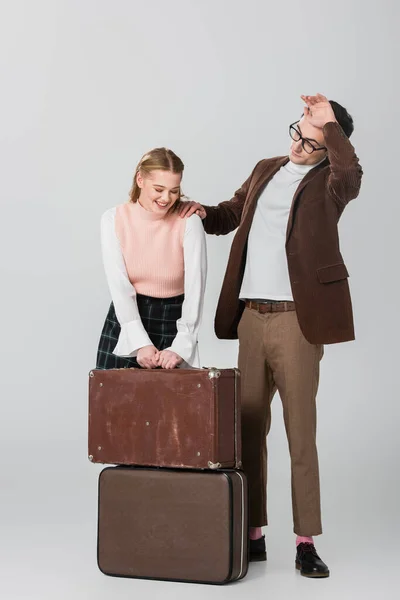 Mujer joven riendo cerca de maletas y novio cansado sobre fondo gris - foto de stock