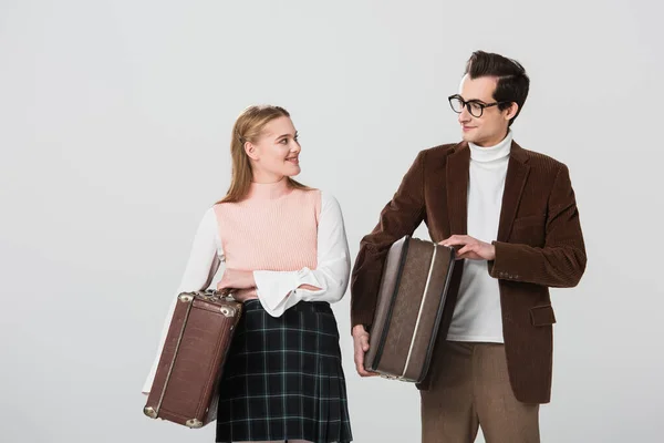 Joven pareja en traje retro sonriendo el uno al otro mientras sostiene maletas aisladas en gris - foto de stock