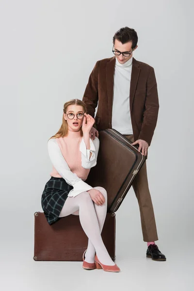 Style rétro femme réglage des lunettes tout en étant assis sur la valise près de l'homme sur fond gris — Photo de stock