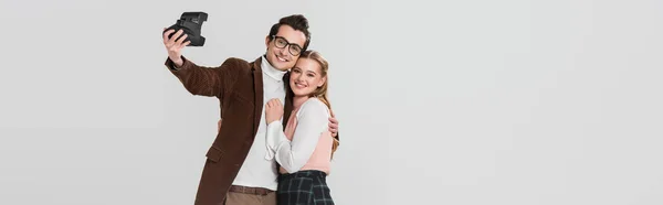 Couple heureux en vêtements rétro prenant selfie sur caméra vintage isolé sur gris, bannière — Photo de stock