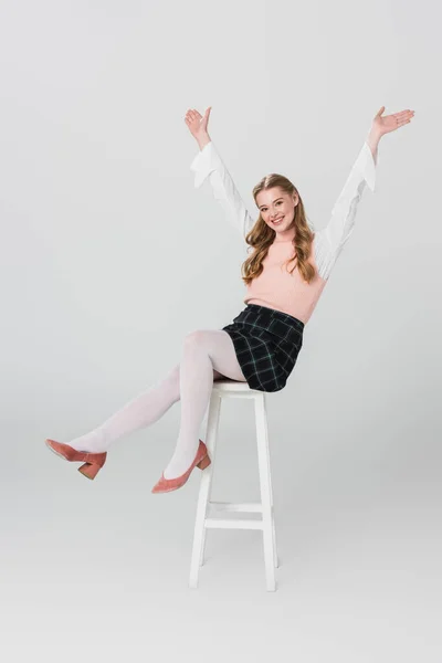 Cheerful woman in vintage outfit sitting on high stool with raised hands on grey — Stock Photo