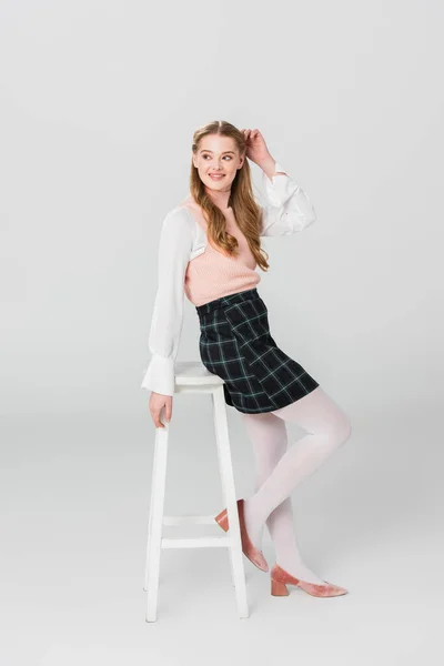 Радостная женщина в винтажной одежде смотрит в сторону, позируя возле высокого стула на сером — стоковое фото