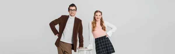 Couple joyeux dans des vêtements rétro posant avec les mains sur les hanches près de haut tabouret isolé sur gris, bannière — Photo de stock