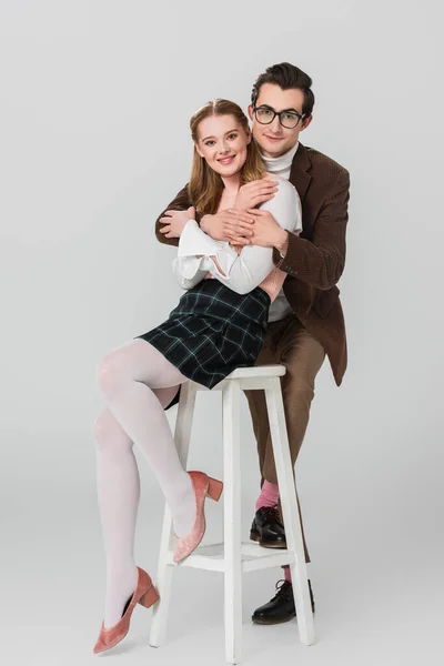 Man in eyeglasses embracing happy woman sitting on high stool on grey background — Stock Photo