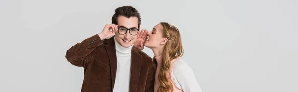 Man in velvet blazer adjusting eyeglasses while girlfriend whispering in his ear isolated on grey, banner — Stock Photo