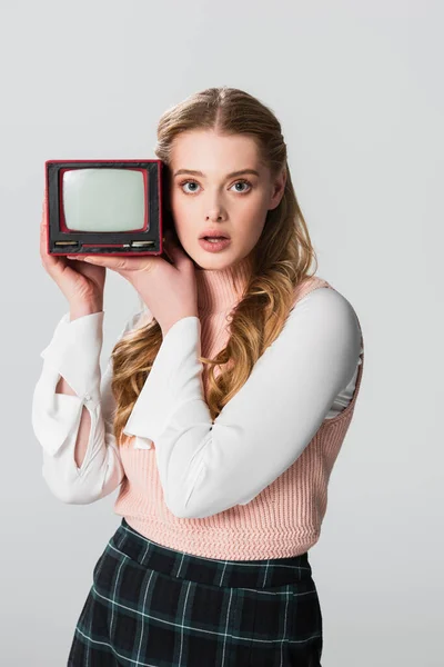 Женщина в стиле ретро смотрит на камеру, держа в руках переносной винтажный телевизор, изолированный на сером — стоковое фото