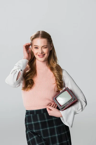 Flirty woman with vintage tv fixing hair while winking at camera isolated on grey — Stock Photo