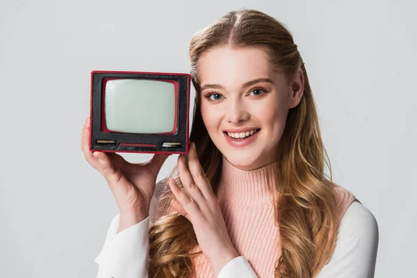 Alegre mujer sonriendo a la cámara mientras sostiene pequeño vintage tv aislado en gris - foto de stock