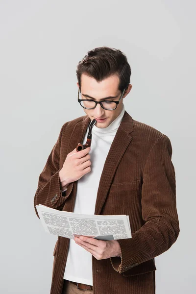 Joven con chaqueta de terciopelo fumar pipa mientras lee periódico aislado en gris - foto de stock