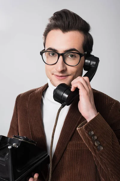 Joven con gafas de vista mirando a la cámara mientras habla por teléfono vintage aislado en gris - foto de stock