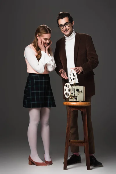 Homme souriant pointant vers projecteur de film près de femme étonnée sur fond gris — Photo de stock