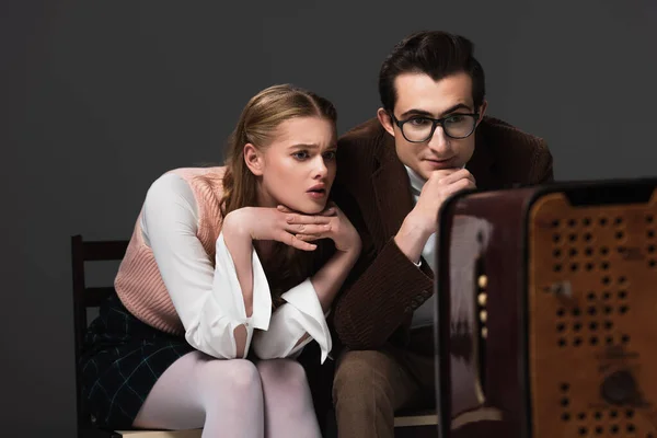 Concentrado casal em roupa vintage assistindo tv isolado em preto, foreground borrado — Fotografia de Stock