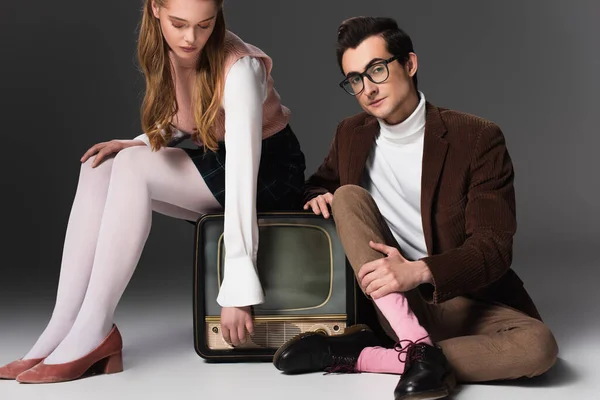 Hombre elegante mirando la cámara cerca de la mujer sentada en la televisión vintage sobre fondo gris - foto de stock