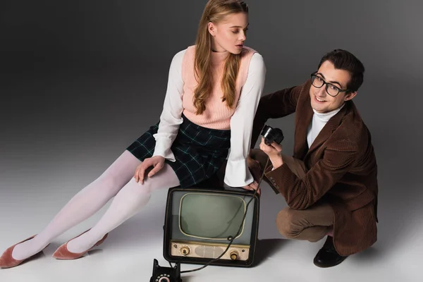 Jolie femme assise sur vintage tv près de sourire homme tenant combiné sur fond gris — Photo de stock