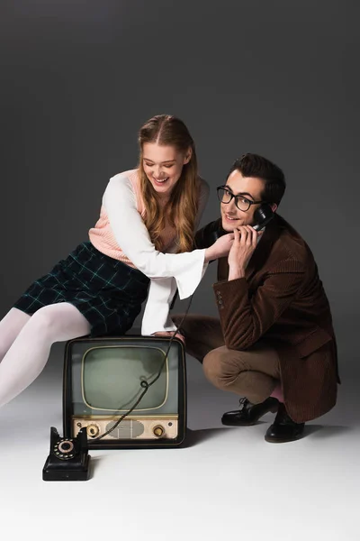 Усміхнений чоловік розмовляє по телефону біля веселої жінки, сидячи на старовинному телевізорі на сірому фоні — стокове фото