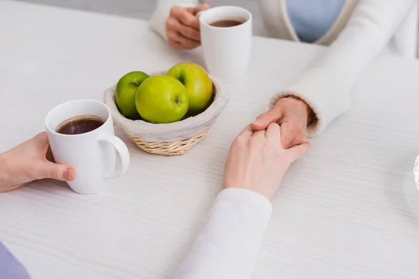 Обрезанный вид пожилой женщины и медсестры, держащихся за руки рядом со свежими яблоками и чашками чая — стоковое фото